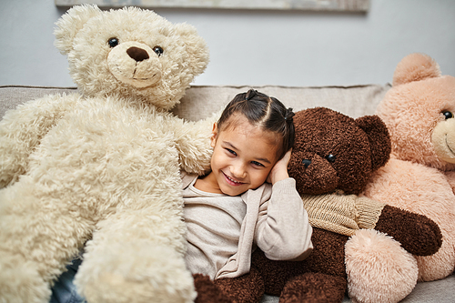 cheerful elementary age girl sitting among soft teddy bears on couch in modern living room, at home