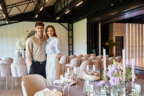 pleased woman pointing at festive table with wedding decor near happy boyfriend in event hall