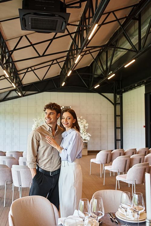 cheerful woman embracing boyfriend and looking at festive table in banquet hall with wedding decor