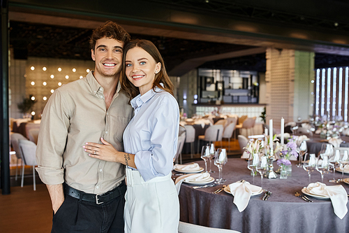 happy couple in love looking at camera in banquet hall with decorated festive tables, special day