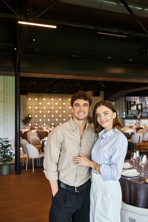 cheerful enamored couple smiling at camera in event hall with festive tables, wedding preparation