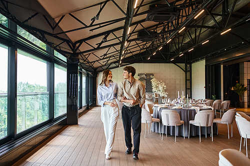 cheerful couple walking along banquet hall with festive tables and wedding decor, romantic setting