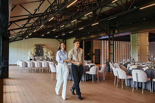 joyful couple walking along festive tables in modern spacious banquet hall with wedding decor