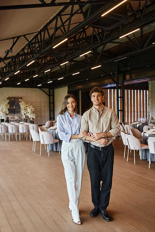 smiling couple in love looking at camera in modern banquet hall with decorated festive tables