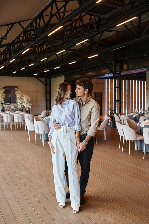 joyful man embracing girlfriend in spacious banquet hall with festive tables and wedding decor