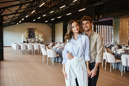 delighted enamored couple looking at camera in modern wedding venue with decorated festive tables