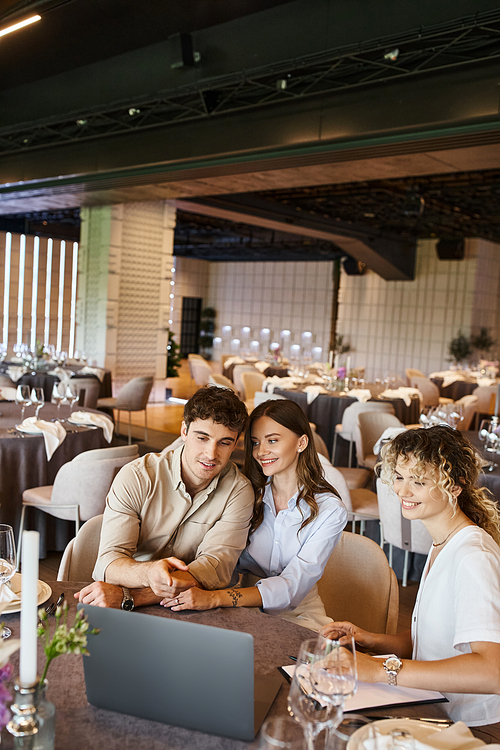 smiling man pointing at laptop near girlfriend and event manager at festive table in wedding venue