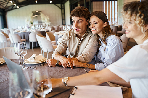 amazed and happy couple looking at laptop near event organizer at festive table, bridal preparation