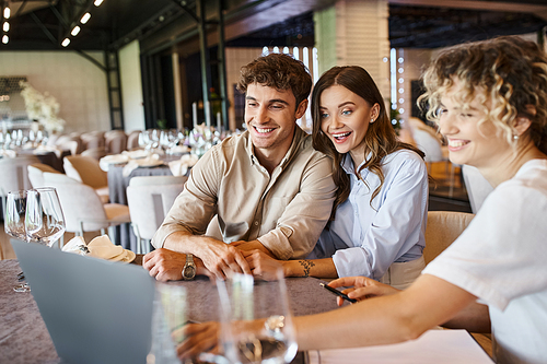 impressed couple looking at laptop near banquet organizer at festive table in wedding venue