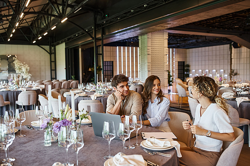 happy couple and professional event manager talking near laptop at festive table in wedding venue