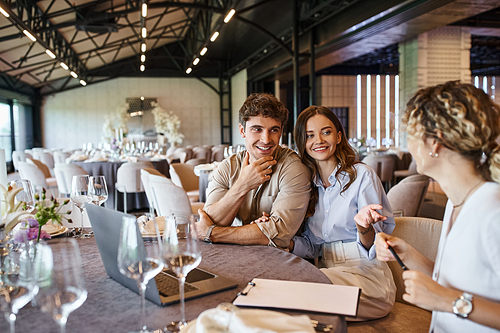 happy couple and event manager discussing wedding preparation at festive table in banquet hall