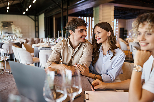 happy couple talking near blurred event manager near laptop on festive table in banquet hall