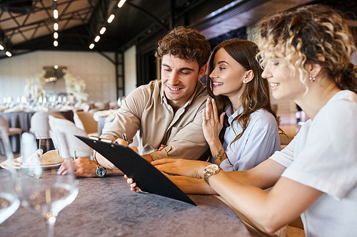 smiling man signing contract near joyful girlfriend and wedding coordinator in modern banquet hall