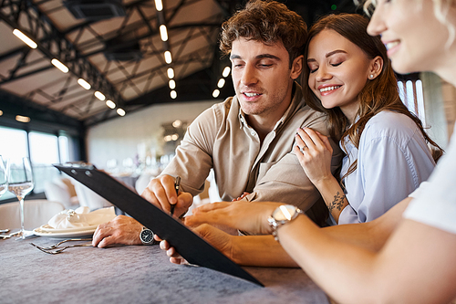 happy man signing contract near young girlfriend and banquet organizer in decorated wedding venue