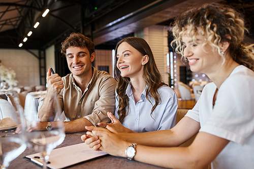 smiling man holding pen near girlfriend and event manager sitting at festive table in wedding venue