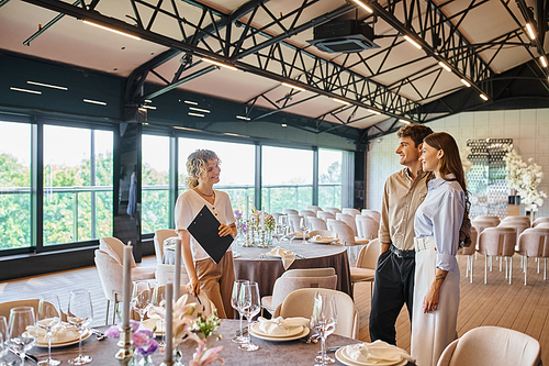 smiling couple in love talking to event decorator near festive tables in modern wedding venue