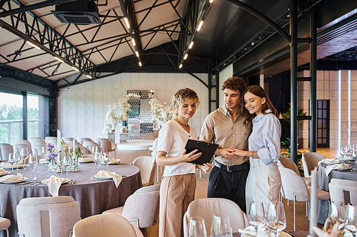 happy couple looking at contract on clipboard near banquet coordinator in decorated event hall