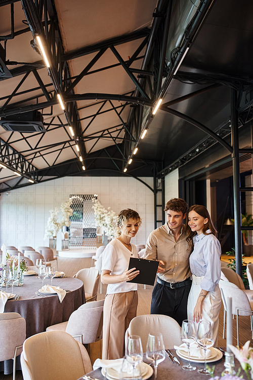 joyful couple looking at contract on clipboard near banquet manager in decorated event hall