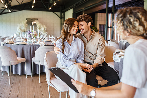 happy enamored couple sitting with closed eyes near event manager with clipboard in wedding venue