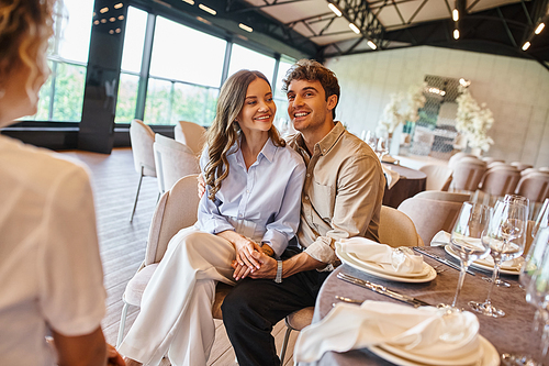 cheerful couple in love sitting at festive table next to event coordinator in wedding venue