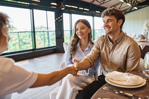 happy man shaking hands with event coordinator near pleased girlfriend in modern wedding venue