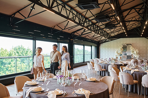 smiling banquet coordinator showing festive tables to joyful couple in decorated wedding hall