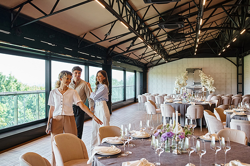 smiling event organizer pointing at festive tables near happy couple in decorated wedding hall