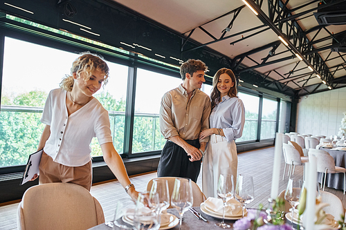 happy couple in love near event manager and tables with festive setting in modern banquet hall