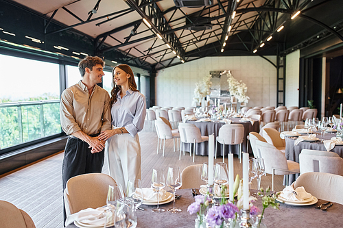 happy couple in love looking at each other near festive table in event hall, bridal preparation