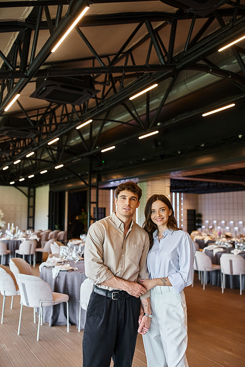 joyful couple in love looking at camera near festive table in event hall, wedding preparation