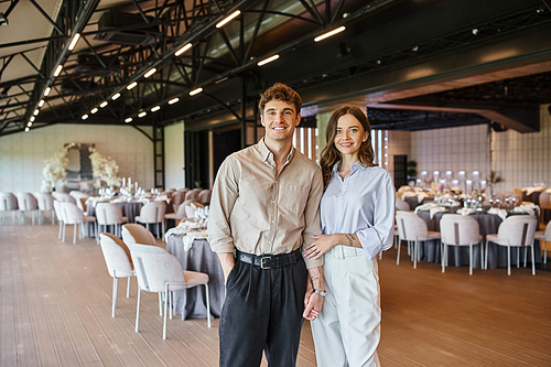 cheerful enamored couple smiling at camera near festive table in wedding venue, romantic setting