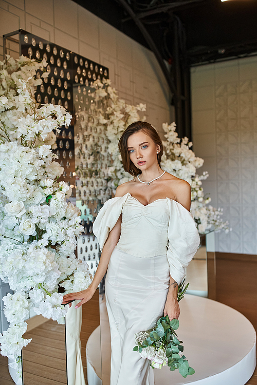 charming woman in white wedding dress looking at camera near floral composition in event hall