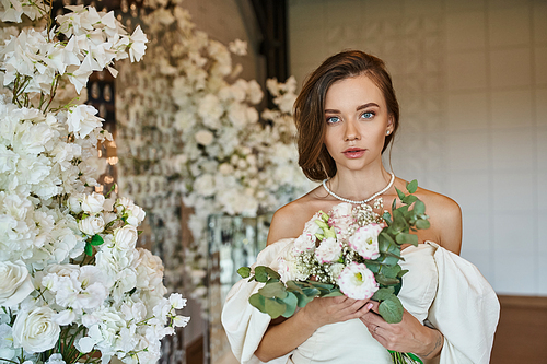 elegant woman in white wedding dress with bridal bouquet looking at camera near festive floral decor