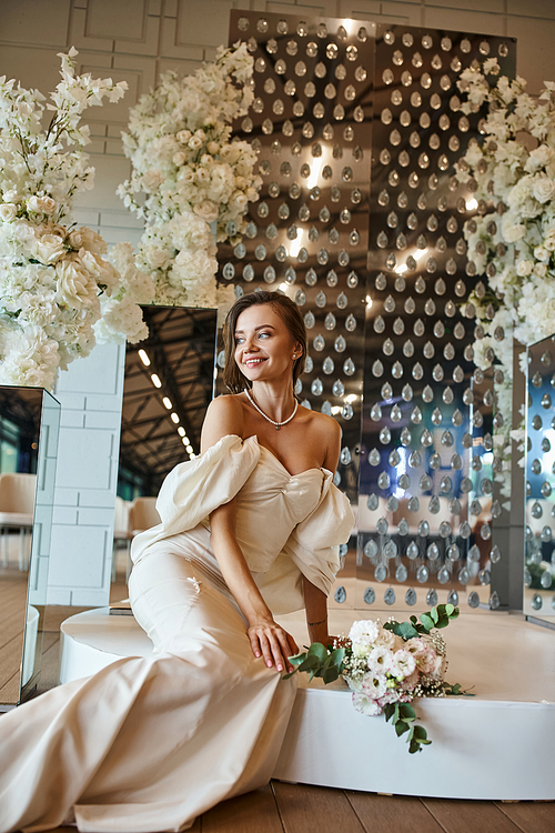 cheerful young bride in white wedding dress sitting near white floral decor in celebration hall