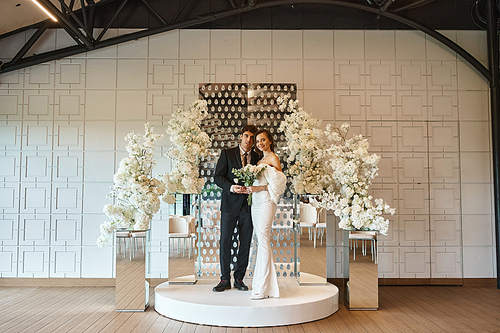 full length of romantic newlywed couple posing in event hall decorated with white blooming flowers