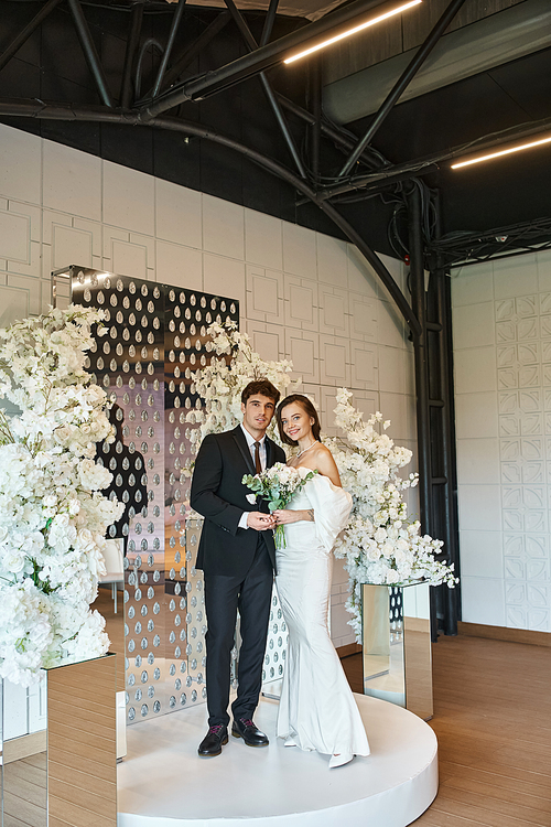 full length of romantic newlywed couple posing in event hall decorated with white blooming flowers