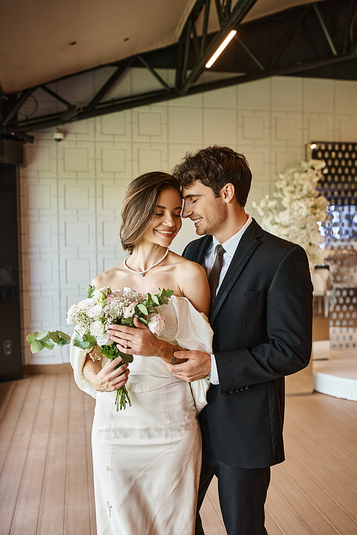 happy and charming bride with wedding bouquet smiling near elegant groom in decorated event hall