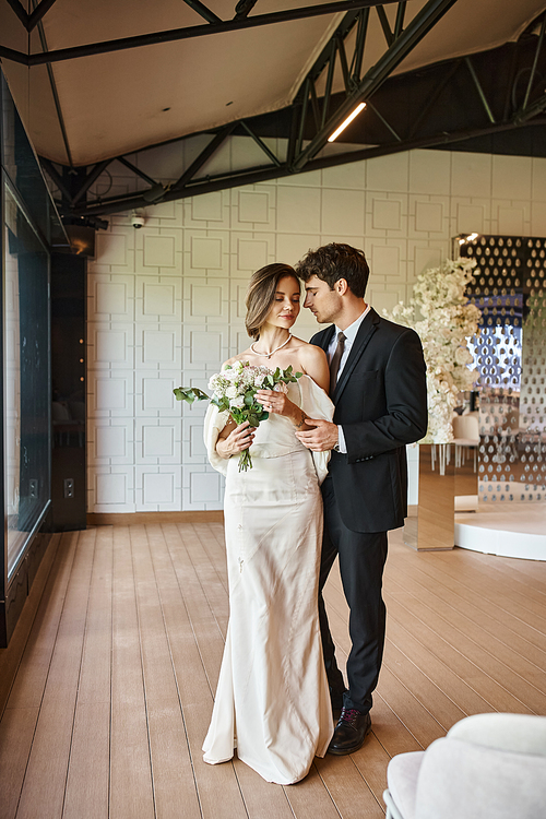 full length of young elegant couple in wedding attire in modern banquet hall with floral decor