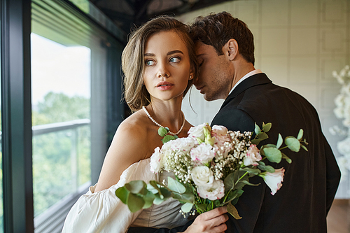 charming bride with wedding bouquet looking away near groom in  black suit in banquet hall