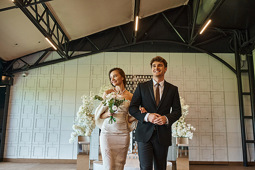 cheerful elegant couple in wedding attire walking in modern event hall with white floral decor