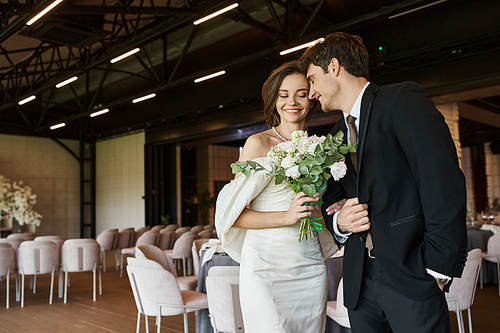 happy groom smiling with closed eyes near charming bride with wedding bouquet in modern banquet hall