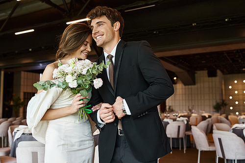 overjoyed groom smiling near young charming bride with wedding bouquet in modern banquet hall