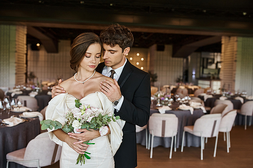 elegant groom with closed eyes embracing charming bride with wedding bouquet in modern event hall