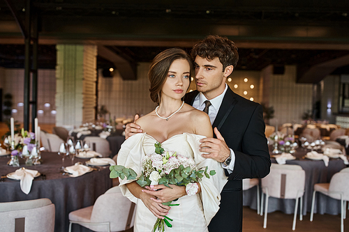 elegant groom hugging young and charming bride with wedding bouquet in modern banquet hall