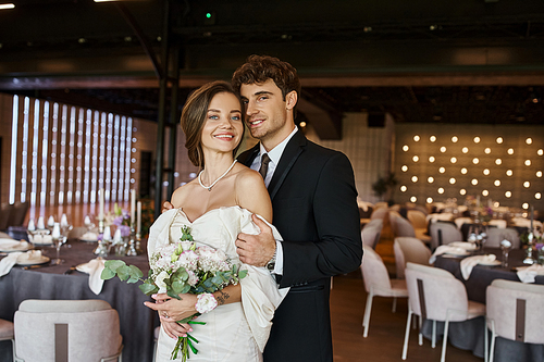 happy young couple in elegant wedding attire looking at camera in decorated celebration hall