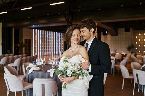 delighted couple in elegant wedding attire smiling at camera in modern banquet hall, banner