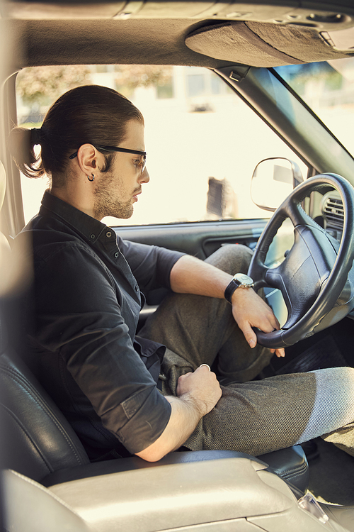 pensive stylish male model with accessories in black outfit behind steering wheel, sexy driver