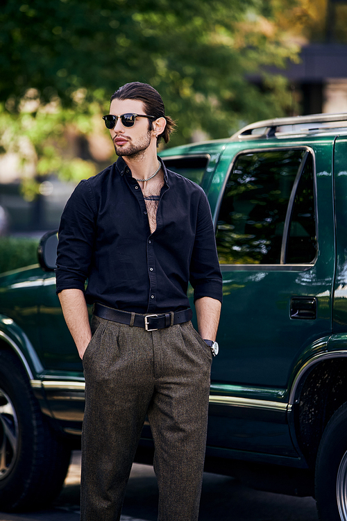 sexy man with accessories in stylish attire standing next to his car with hands in pockets