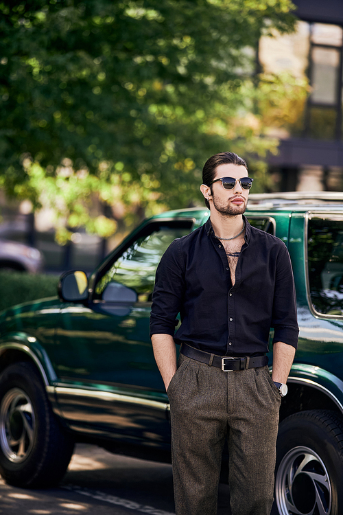 vertical shot of charming man in elegant black attire posing next to car with hands in pockets