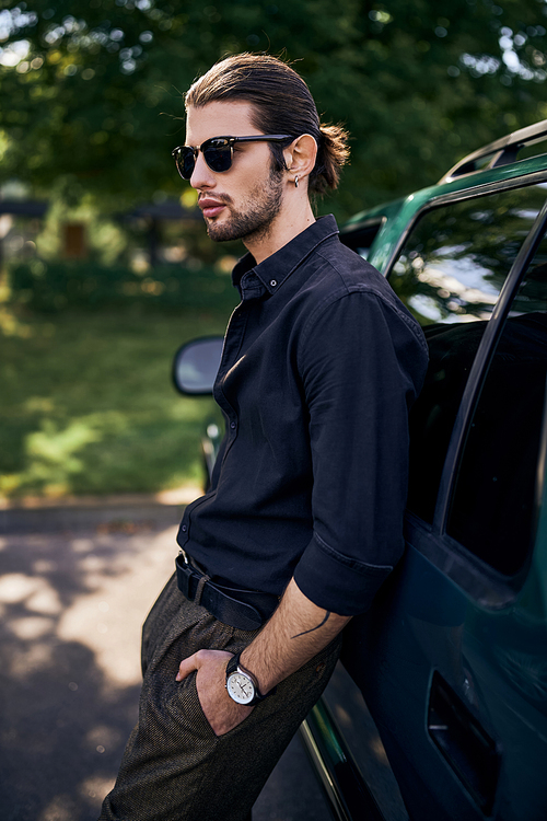 vertical shot of good looking young man in black shirt with ponytail posing near car, sexy driver